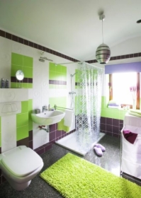 Jasna łazienka po remoncie z zielono-brązowymi akcentami: płytki na ścianach, dywanik, zasłona prysznicowa, umywalka z baterią, toaleta i okno z fioletową zasłoną.